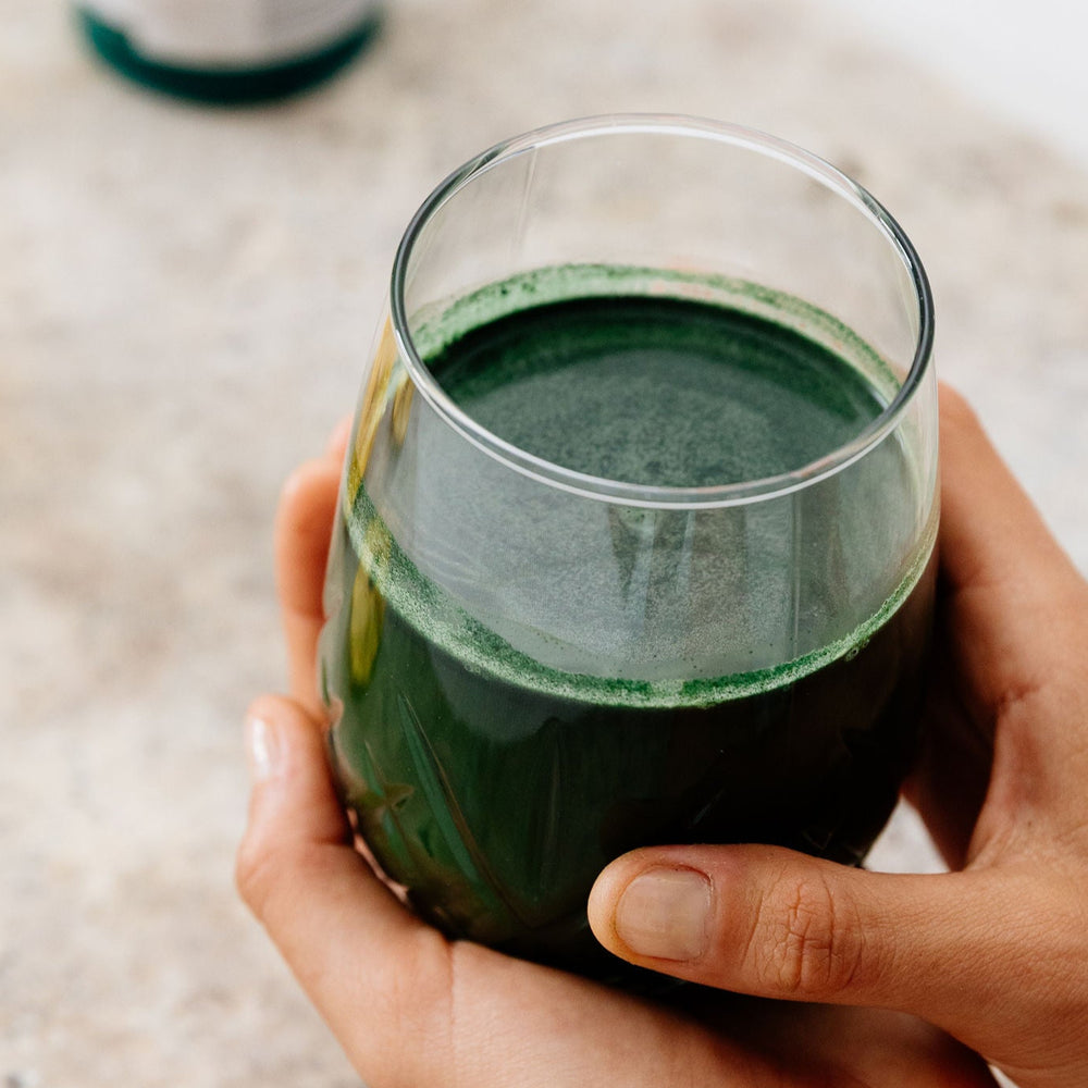 Person holding a glass of Phytality Phytoplankton Super Greens with a bottle and dried fruit on a light surface
